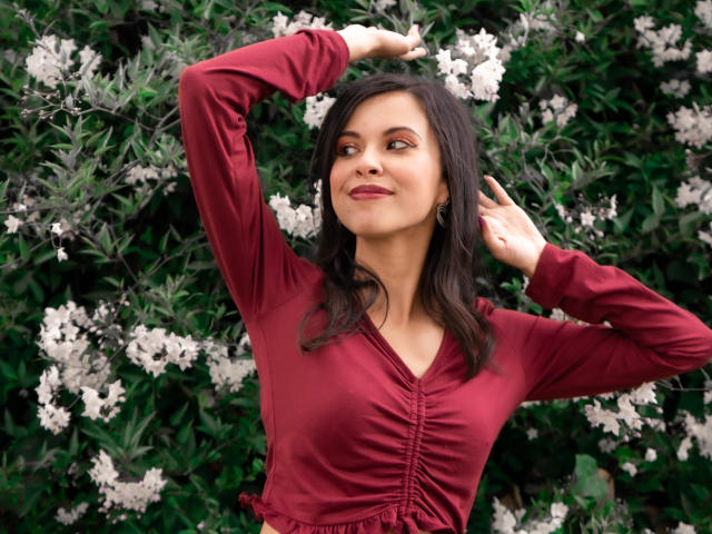 TaylaCooperk with dark hair, wearing a red dress, arms raised, hands in hair, smiling, white flowers in greenery behind her, medium tits, earrings, subtle makeup.