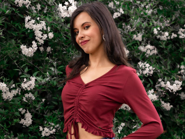 TaylaCooperk with medium tits, wearing a red crop top, standing in front of white flowers, has long dark hair, and a slight smile, hands on hips, looking to the side.