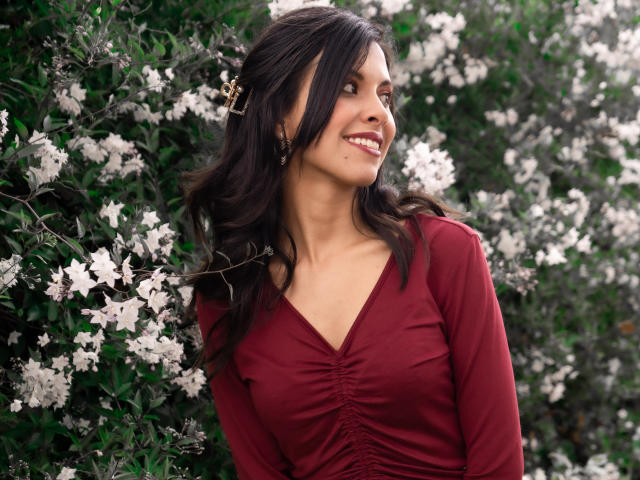 TaylaCooperk with wavy dark hair, red dress, small tits, smiling, looking to the side, white flowers in background, green leaves, brown earrings, standing in garden.