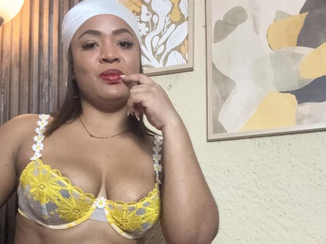 AliceBooty with brown skin, wearing a white headscarf, yellow and white lace bra, and red lipstick, touching her lip with her finger. Boobs are visible, she's looking at the camera.