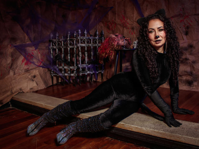 SamanthaAntolin is in a dark, velvety cat costume with black gloves and boots, lying on a wooden floor. She has curly black hair, dark makeup, and cat ears. Her boots are sparkly with a blue hue. In the background, a metal rack with a red, bloody object is on a brown, textured wall with purple graffiti. The lighting is dark and moody. The image has a gothic, edgy vibe.