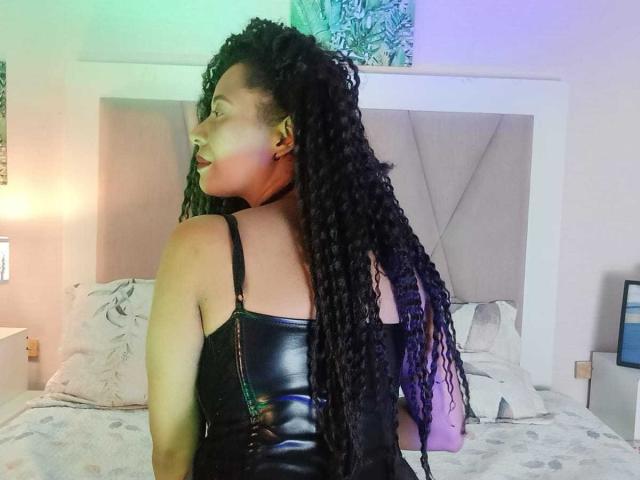 KenyaKarath with long black braids, wearing a black leather dress, showing her back and looking over her shoulder, in a bedroom with a grey upholstered headboard.