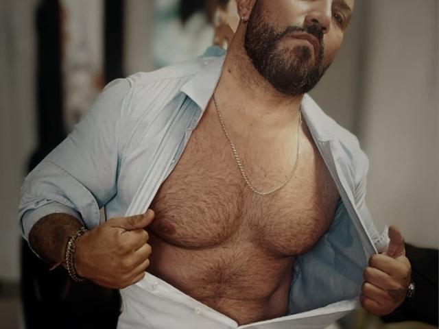 BardoCkroy69 with a beard, chest hair, and a blue shirt open, showing his hairy, muscular chest and a silver necklace, holding the shirt with both hands.