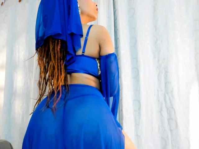 KendallQuinn in a blue sheer outfit, showing her back with visible ass, long brown wavy hair, and off-shoulder bra, standing in front of white curtains.