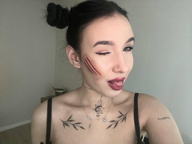 NikkiGoldX with black hair in a bun, red scratch marks on cheek, dark lipstick, tattoos on chest and neck, black strap top, winking, tongue out, light skin.