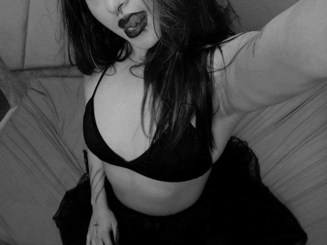 Black-and-white photo of SamaraCuttie. She has long, dark hair, a nose ring, and dark lipstick. She's wearing a black bra that reveals her mid-sized breasts and a dark, semi-sheer skirt. She's holding a selfie stick, looking down at the camera. Her right arm is partially visible. The texture of the bra is slightly textured. Her skin is light. The image has a slightly rough, intimate feel.