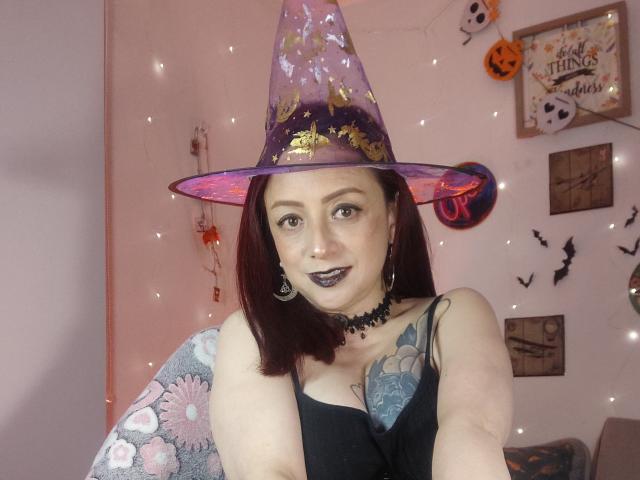 CatalinaMonyque with dark lipstick, wearing a purple witch hat, black choker, and black top, showing her tattooed tits and smiling at the camera. Red hair, pink walls.