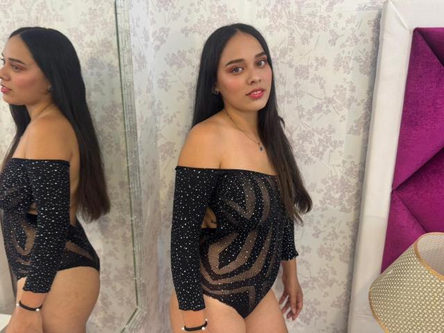 BonnieJaz está de pie frente a un espejo, vestida con un body negro transparente con lunares, que deja ver sus pequeños pechos y su zona púbica a través de la tela..
