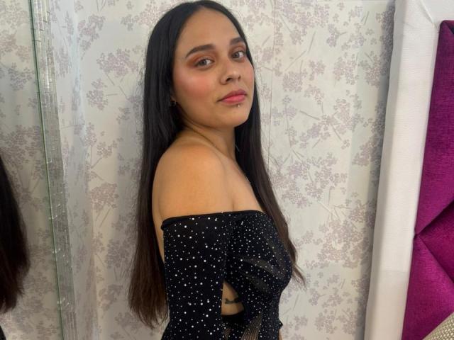 BonnieJaz stands facing slightly to the right, wearing an off-the-shoulder black dress with small white sparkles. She has long, straight black hair, brown eyes, and pink lipstick. Her skin is medium brown. The background features a floral-patterned wallpaper. To the right, a purple fabric is partially visible. The image is a clear photograph with natural lighting. BonnieJaz's expression is neutral, with a slight, closed-mouth smile.