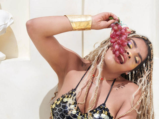 LexieBloom, a black woman with blonde braids, holds a cluster of purple grapes to her mouth. She wears a black sequined bikini top with gold accents, and a gold bracelet. Her eyes are closed, and she has yellow eye makeup. She has a tattoo on her right shoulder. The background is a simple white wall. The image is bright and focused on LexieBloom. The grapes are in her right hand. The texture of her braids contrasts with the sequined top.