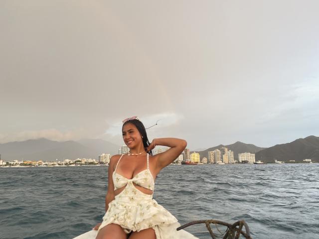 SarahPrincess with tan skin, black hair, and small breasts in a white floral dress, sitting on a boat, water behind, mountains and city in the background, smiling.