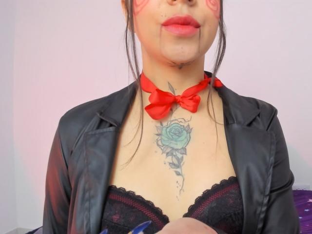 KayleeM with red lipstick, black lace bra, and red bowtie, wearing a black leather jacket, has a blue rose tattoo on her chest, holding a blue pen.
