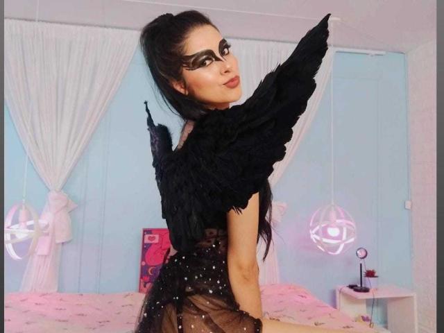 AmberLenox with black feather wings, black mask, and sheer black dress, sitting on pink bed, black hair in ponytail, looking back, bedroom with pink and white decor.