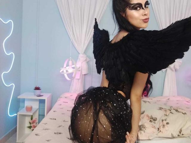 AmberLenox with black wings, wearing a sheer black dress with sparkling details, showing her round ass, looking back with dark makeup and black hair, in a light blue room.