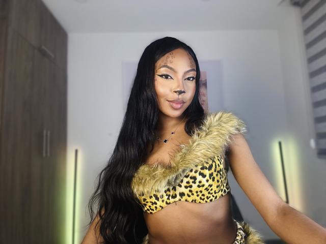 MissMaary with long black hair, wearing a leopard print bra with furry shoulder straps, showing her small tits and a confident smile. She's standing in a brightly lit room.