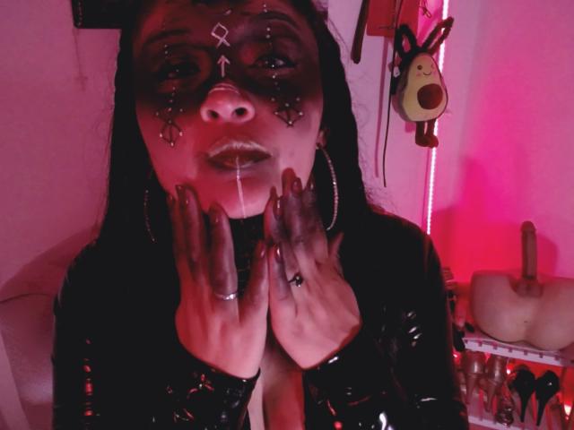 LindaSophieX with black mask, red lips, and long black hair, wearing shiny black latex, hands near face, red and pink lighting, dildo and mask on shelf in background.