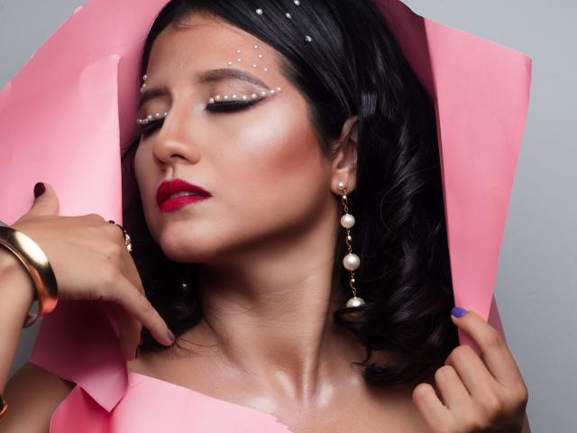 RenataRivas met zwart haar, roze papier, parel oorbellen, glinsterende eyeliner, rode lipstick, een gouden armband en blauwe nagellak, met gesloten ogen, hand die haar wang aanraakt. Borsten, tieten, sexy..