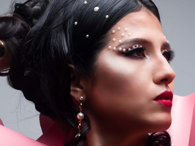 RenataRivas has dark hair, pearl dots on her forehead, and red lipstick. She's wearing pearl earrings and a pink garment. Her eyes are brown and she's looking to the side.