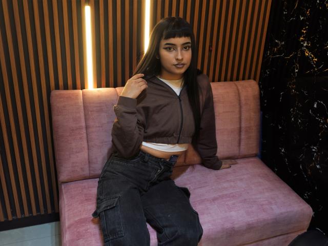 CleoRosee with dark hair, brown jacket, white crop top, black jeans, sitting on pink velvet sofa, hand on jacket, looking at camera, medium skin, full lips, subtle makeup.