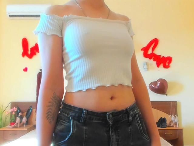 Steisii vestindo uma blusa branca listrada com cava caída, mostrando seus seios, com uma tatuagem no braço direito, e calças jeans pretas, em pé em um quarto com decorações vermelhas com a palavra "amor"..