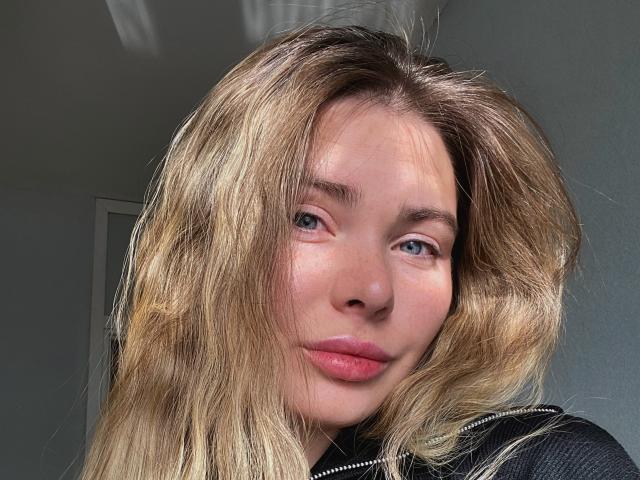 KattyMonica has blonde wavy hair, blue eyes, and pinkish lips. She's wearing a black zip-up. Her face is slightly flushed. She looks cute and sexy.