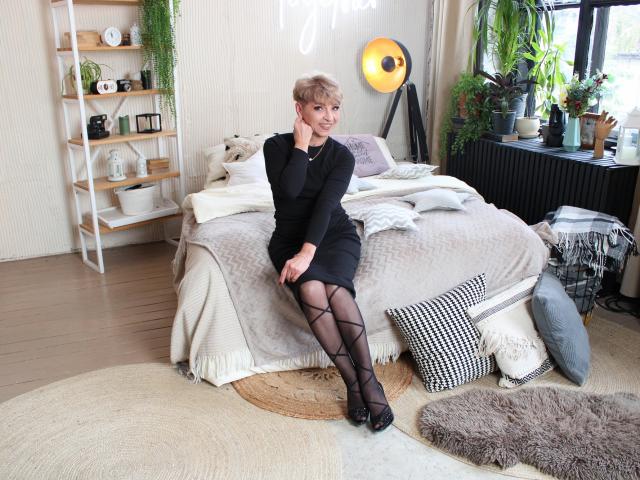 EllaBonito, blonde, black dress, black lace-up stockings, black heels, sitting on bed, hands on knee, smiling, bed with gray blanket, white pillows, wooden floor, plants, modern room.
