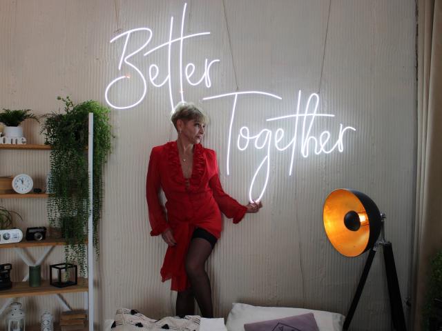 EllaBonito, short blonde hair, red ruffled dress, black stockings, standing, touching neon "Better Together" sign, looking right, next to orange floor lamp, indoor setting.