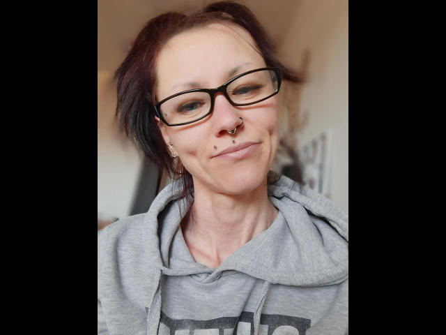 Izabella-mit-Z-hot has dark hair, black glasses, a nose ring, and two small facial piercings. She's wearing a gray hoodie with a subtle smile.