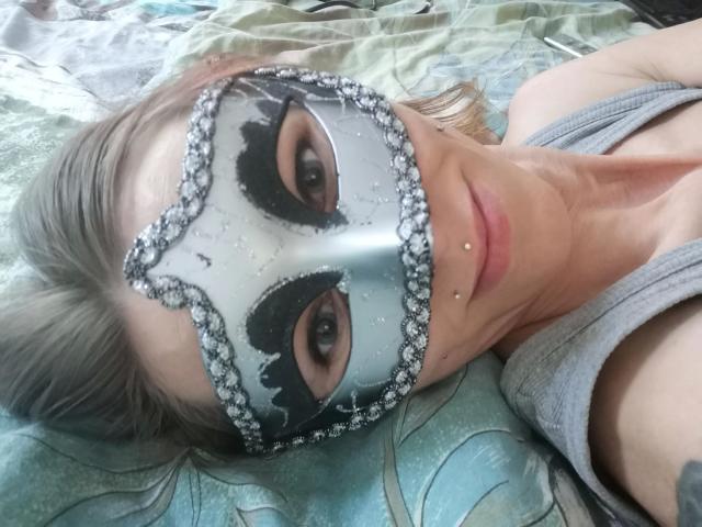 Izabella-mit-Z-hot lying on a bed, wearing a sparkly blue and black mask, with gray hair, gray tank top, and facial piercings, showing a sexy, relaxed expression.