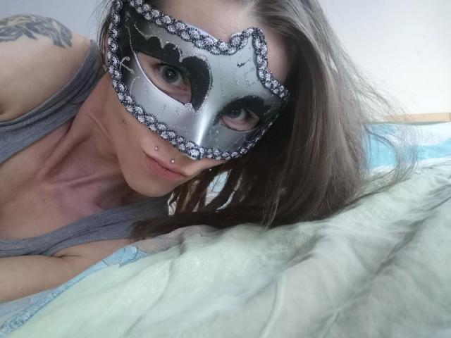 Izabella-mit-Z-hot with a silver masquerade mask adorned in diamonds, wearing a gray tank top, lying on a bed with green and blue sheets, sporting a nose piercing.
