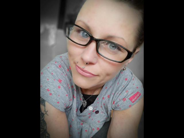 Izabella-mit-Z-hot has blue eyes, black glasses, nose piercings, and a septum piercing. She's wearing a gray shirt with pink patterns, and has tattoos on her arms.