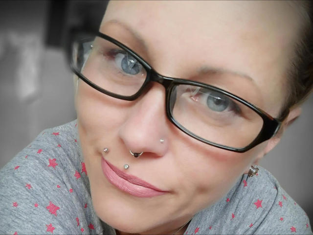 Izabella-mit-Z-hot with glasses, nose piercings, lip piercing, and pink lipstick, wearing a grey shirt with red patterns, has blue eyes and a subtle smile.