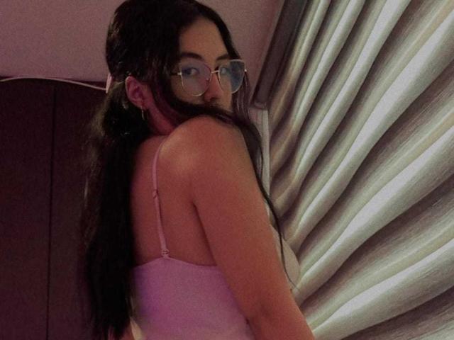 MiaDiiaz with long black hair, glasses, pink tank top, showing her bare back and tits, looking over her shoulder, in a dimly lit room.