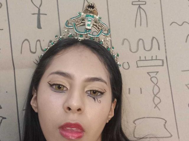 SoffiRousee's face is centered in the image. She has medium brown skin, dark straight hair, and wears a jeweled crown with green and gold accents. Her brown eyes have black eyeliner with a small black tattoo-like mark under her right eye. She has pink lipstick and a slight, neutral expression. The background is a beige wall with black, abstract line drawings. The crown's intricate design contrasts with her smooth, unadorned face. The image focuses on SoffiRousee's face and the decorative crown.