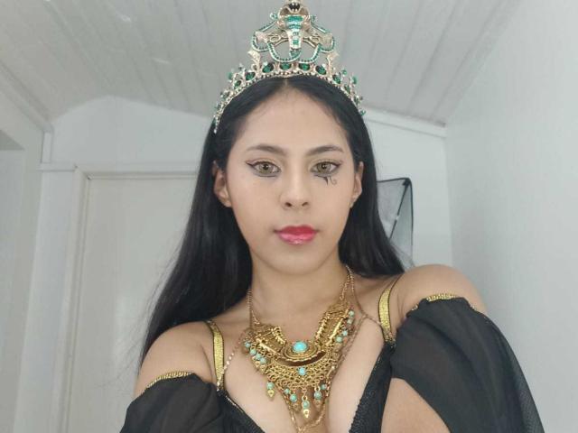 SoffiRousee with long black hair, wearing a green and gold tiara, black off-shoulder dress, and ornate necklace, showing small tits, with pink lipstick and subtle makeup.