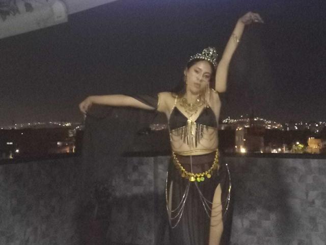 SoffiRousee dancing at night, wearing a black bikini top with gold chains, brown skirt with gold ornaments, and a jeweled headpiece, arms raised, city lights in the background.
