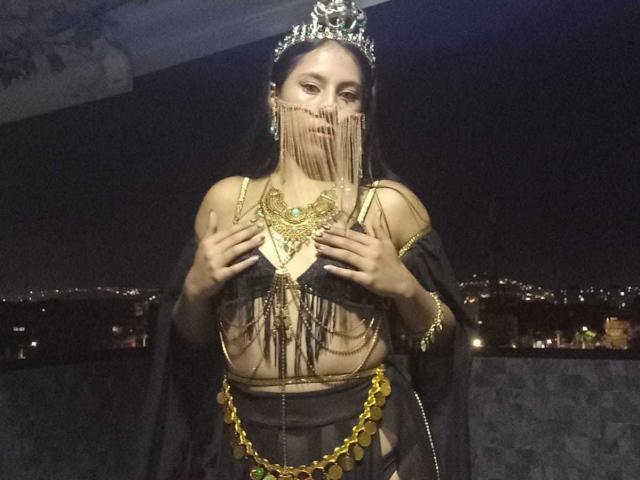 SoffiRousee with dark hair, wearing a jeweled tiara, chain lingerie, and sheer black veil, holding gold necklace, exposing her tits, against a night-time city skyline.