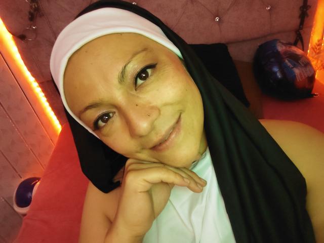 SusanaHotSex with a nun's habit, smiling, has a nose ring and is resting her chin on her hand, showing her bare shoulders and a slight smile.