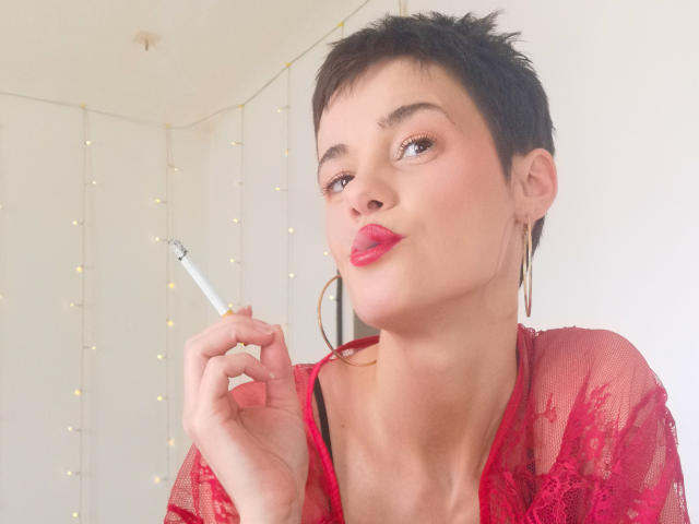 LaPhoenixElo with short black hair, red lace shirt, and pink lipstick, holding a cigarette, looking up with a pouty expression, wearing gold hoop earrings.