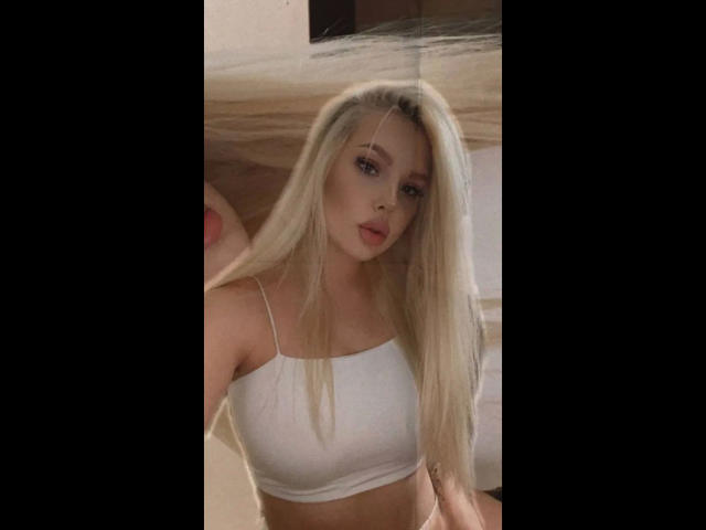 Blonde Byanka-hot with long hair, wearing a white crop top, showing her tits, pink lips, and light makeup, taking a selfie in a softly lit room.