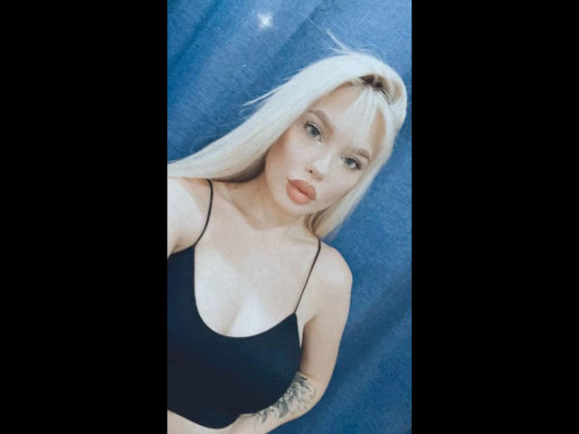 Blonde Byanka-hot with blue eyes, wearing a black sports bra, showing small tits and a tattoo on her arm, has red lipstick, and a slightly pouty expression.