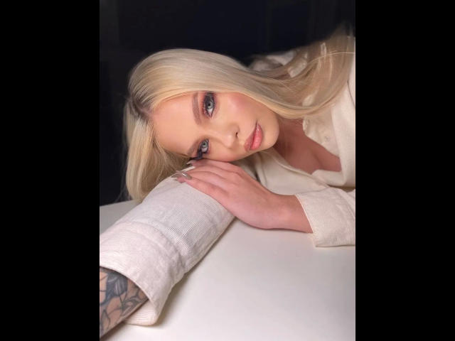 Byanka-hot with blonde hair and blue eyes, leaning on a white surface, wearing a white shirt, showing tattooed arm, looking seductively at camera.