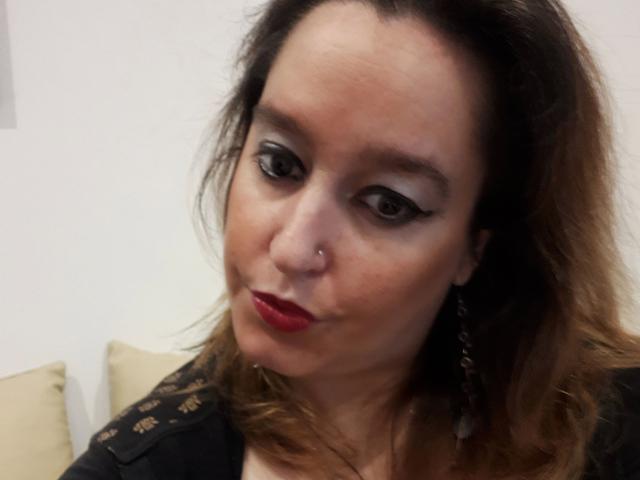 AnitaRed-hot hat eine mittelbraune Hautfarbe, dunklen Eyeliner, roten Lippenstift, eine Nasenring und welliges braunes Haar. Sie trägt eine schwarze Spitzenbluse mit hängenden Ohrringen und blickt leicht nach unten..