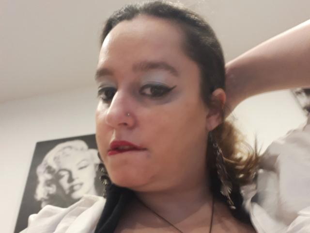 AnitaRed-hot heeft donker haar, blauwe oogschaduw, rode lippenstift, een neusring, en kijkt naar beneden met haar hand achter haar hoofd, en draagt een witte top..