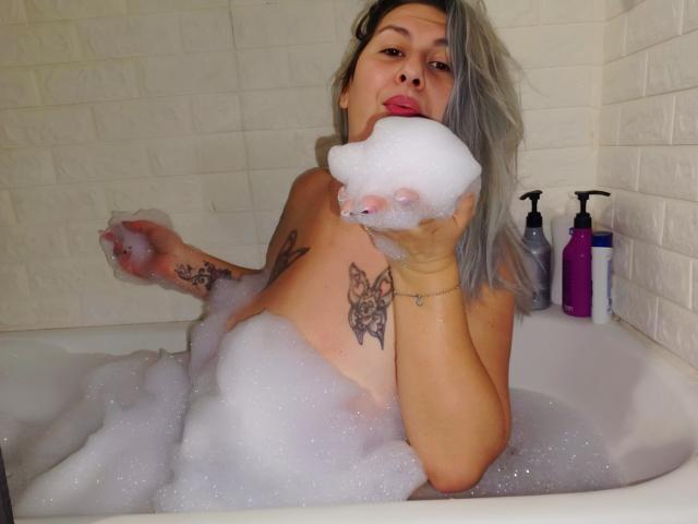 LilithGreenn, naked, with foamy bubbles covering her tits and ass, holding a soapy puff to her mouth, has tattoos on her arm and side, in a white-tiled bathtub.