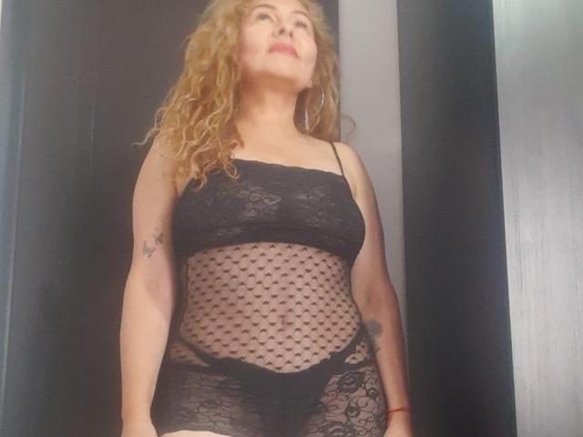 MiaAmoretti with curly red hair, wearing black lace lingerie with a mesh belly, standing, looking up, visible tattoo on right arm, red bracelet on left wrist.