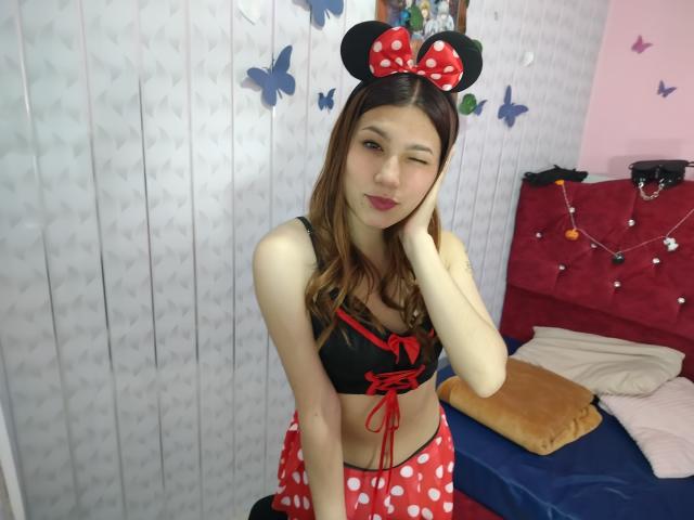 CeelesteBonnett with brown hair, winking, wearing a black crop top with red lace, red and white polka dot skirt, and Minnie Mouse headband, in a bedroom, smiling.
