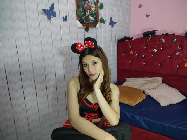 CeelesteBonnett with brown hair, wearing black and red Minnie Mouse outfit, headband with bow, black top, red skirt, hand on cheek, sitting on black suitcase, in bedroom.