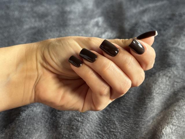 AquaLady's hand with shiny black, almond-shaped nails, slightly curled fingers, on a gray, textured surface. Her skin is light, and the nails are polished and glossy.