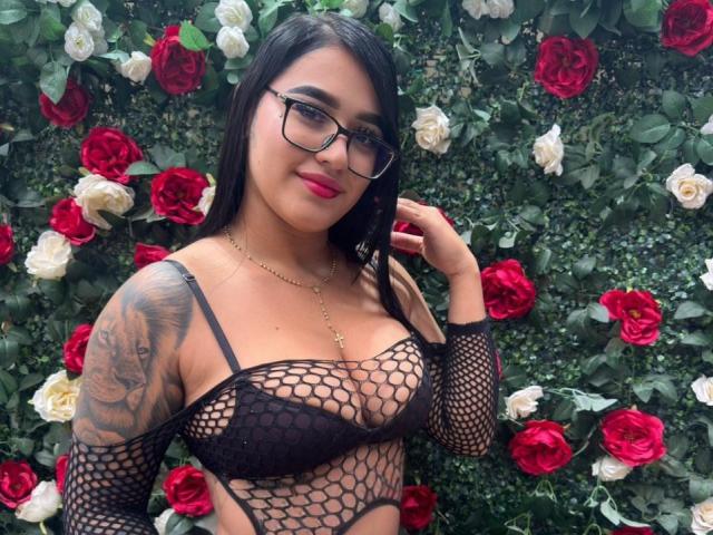 MelodyRuso trägt ein schwarzes Netzoberteil, rote Lippen, eine Brille und hat einen tätowierten Arm. Sie zeigt ihre Brüste vor einer Wand aus roten und weißen Rosen. Sie berührt ihr Haar und lächelt..